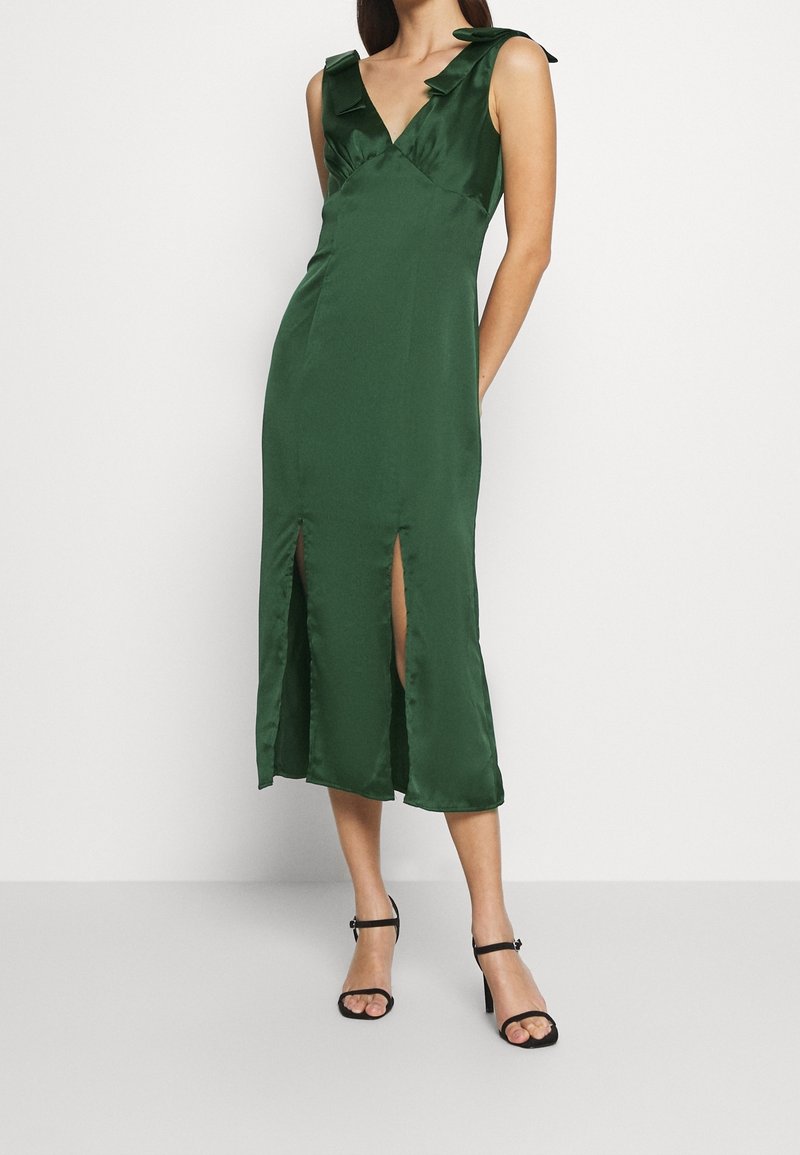 Femme portant une robe midi sans manches vert foncé avec un décolleté en V, des nœuds aux épaules et deux fentes à l'avant, associée à des talons hauts noirs à lanières.