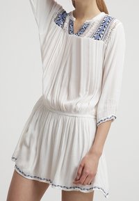 Vit klänning med en texturerad tyg, med blå blommig broderi på halsringningen och fållen. Elastisk midja, lös passform och långa ärmar.