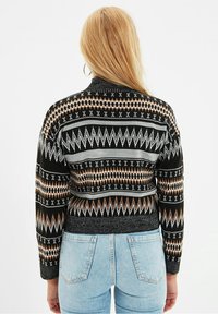 Schwarzer Pullover mit geometrischen Mustern in Grau, Braun und Kupferakzenten. Verfügt über einen hohen Kragen und Rippbündchen. Getragen mit hellblauen Jeans.