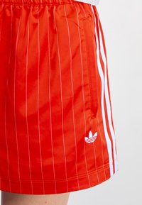 Röda sportshorts med vita vertikala ränder, elastisk midja, sidoficka och en liten vit Adidas-logga på nedre vänstra sidan.