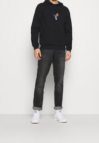 Schwarzer Hoodie mit farbenfrohem Kolibri-Stickerei, kombiniert mit dunklen Jeans und weißen Sneakern mit einem Logo. Glattes Material und lockere Passform.