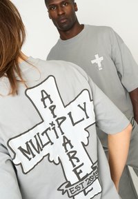 Las camisetas de gran tamaño en color gris presentan un gran gráfico de una cruz y texto en la parte posterior, con un logotipo más pequeño en el frontal. Textura de tela suave.