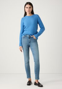 Blauer Strickpullover mit langen Ärmeln, kombiniert mit hellblauen Slim-Fit-Jeans und dunkelbraunen Loafers. Einfaches, körpernahes Design mit gerippten Akzenten.