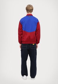 Giacca a vento rossa e blu con orlo elasticizzato, caratterizzata da un design a blocchi di colore a contrasto. Indossata con pantaloni scuri e scarpe bianche.