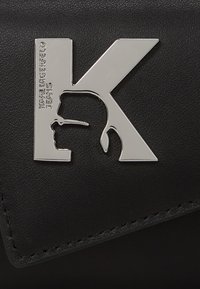 Černá umělá kůže s metalicky stříbrným logem K ve tvaru profilového siluety, vedle textu "KARL LAGERFELD JEANS." Texturovaný povrch.