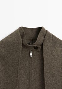 Cappotto in lana infeltrita di colore marrone tenue, con colletto alto, zip anteriore e chiusura a bottone per un dettaglio in più.