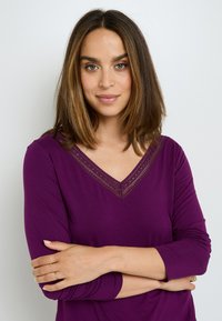 Haut violet à manches longues avec un décolleté en V et des détails en dentelle. Tissu lisse avec une texture douce. Le modèle pose les bras croisés.