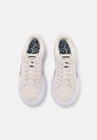 Baskets en daim beige avec une semelle en caoutchouc blanc, présentant des accents noirs, des lacets crème et un intérieur à motifs. Détail de marque doré présent.