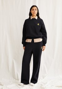 Schwarzer Sweatshirt mit gelbem Schleifenlogo, übereinandergelegt mit einem gestreiften Kragenshirt. Kombiniert mit locker sitzenden schwarzen Hosen. Glatte Stoffstruktur.