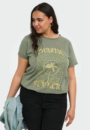 Kvinde med langt mørkt hår iført en grøn "Chasing Summer" t-shirt med palmebladsgrafik, der holder en lys blå denimjakke i venstre hånd.