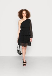 Vestido preto de um ombro só com saia em camadas, textura transparente e cintura elástica. A modelo segura uma pequena bolsa preta e usa sandálias pretas de tiras.