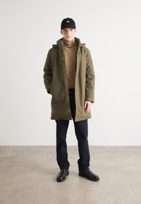 Olivgrüner langer Mantel mit Reißverschluss und Druckknopfverschluss, getragen über einem beigen Rollkragenpullover, kombiniert mit marineblauen Hosen und schwarzen Stiefeln.