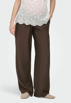 Mujer que lleva pantalones anchos marrón oscuro, camiseta sin mangas blanca bordada y zapatos beige tejidos tipo mocasín, de pie con una mano en el bolsillo.