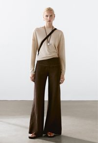 Maglione lavorato a maglia beige abbinato a pantaloni larghi marroni e sandali neri. L'outfit presenta una borsa a tracolla nera e una collana con ciondolo in argento.