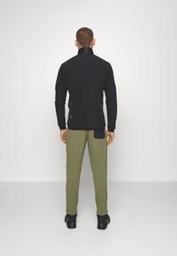 Giacca nera a maniche lunghe con colletto alto e tessuto strutturato, abbinata a pantaloni verde oliva con tasca laterale e finitura liscia.