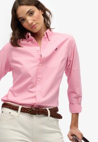 Mujer vestida con una camisa rosa abotonada con un pequeño logo, pantalones blancos y un cinturón trenzado marrón, sosteniendo el asa de un bolso marrón oscuro.