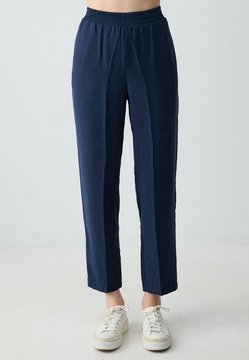 Marineblaue, leichte Hose mit elastischem Bund und geradem Bein. Glatte Textur und dezenter Glanz. Kombiniert mit weißen Sneakers.