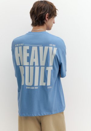 Person med kort brunt hår iført en blå t-shirt med stor off-white tekst "HEAVY BUILT" og beige bukser, vendt væk fra kameraet.
