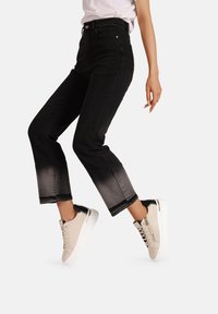 Jeans in denim nero con una sfumatura ombre che degrada verso colori più chiari all'orlo, abbinati a sneakers beige con dettagli neri e suole bianche.