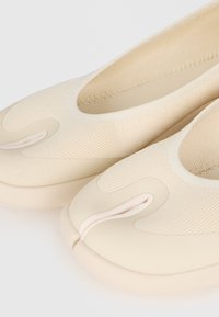 Κοντινή λήψη μπεζ παπουτσιών slip-on με στρογγυλεμένο σχεδιασμό διαχωρισμένου δακτύλου και υφασμάτινο πάνω μέρος με υφή.