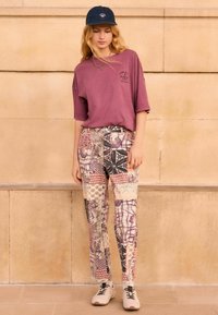 Burgundyröd oversized t-shirt, mönstrade patchwork-byxor i olika färger och beiga sneakers. En enkel keps kompletterar den avslappnade stilen.