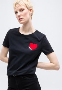 Νέος με κοντά ξανθά μαλλιά και μαύρο μπλουζάκι με κόκκινη καρδιά και την επιγραφή «AMOUR» στο στήθος, σε απλό φόντο.