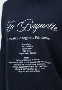 Megztinis su baltu prancūzišku tekstu, kuriame pateikiama bagetės receptas, įskaitant ingredientus ir kepimo etapus, tamsiai mėlynos spalvos fone.