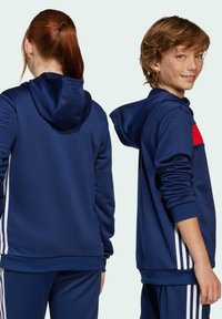 Mornarsko plava dukserica s crvenim akcentom, s prednjim džepom, podesivim kapuljačom i bijelim bočnim prugama. Mekani, strukturni materijal, opušteni kroj.