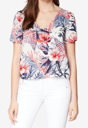 Blouse à manches courtes à motif floral, présentant un motif tropical bleu marine et rose, un col en V et un tissu léger. Associée à un jean blanc.