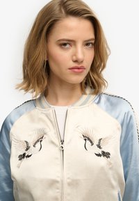Bomber jacket crema con maniche in satin blu e disegni di gru ricamati. Presenta una zip frontale e polsini a coste. Tessuto morbido e silhouette aderente.
