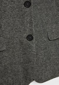 Giacca grigia in misto lana con texture a zigzag, tre chiusure con bottoni neri e due tasche frontali. Design sartoriale con taglio classico.