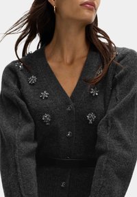 Szary kardigan wykonany z miękkiej wełny, z dekoltem w serek, bufiastymi rękawami, ozdobiony kryształowymi detalami kwiatowymi oraz czarnym paskiem w talii.