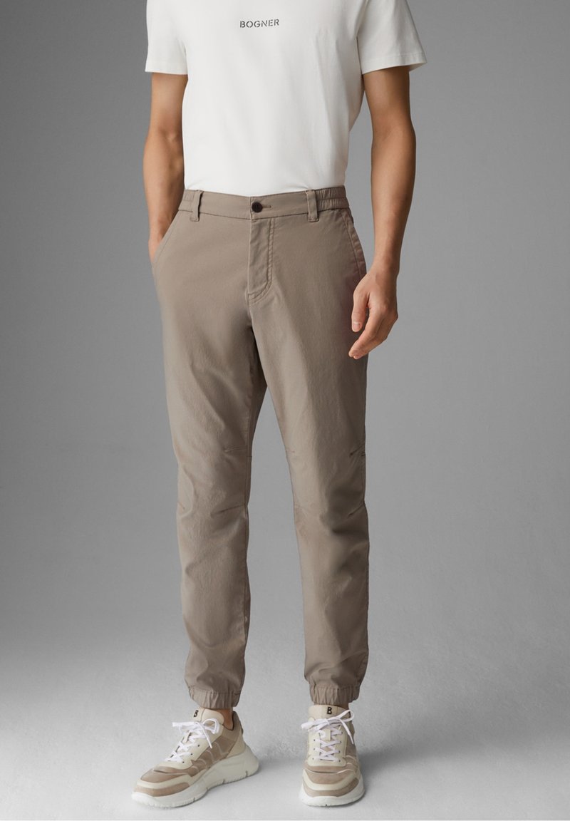 Homme portant un pantalon de jogging beige avec des poignets élastiques, des baskets blanches et un t-shirt blanc avec le texte "BOGNER" sur un fond gris.