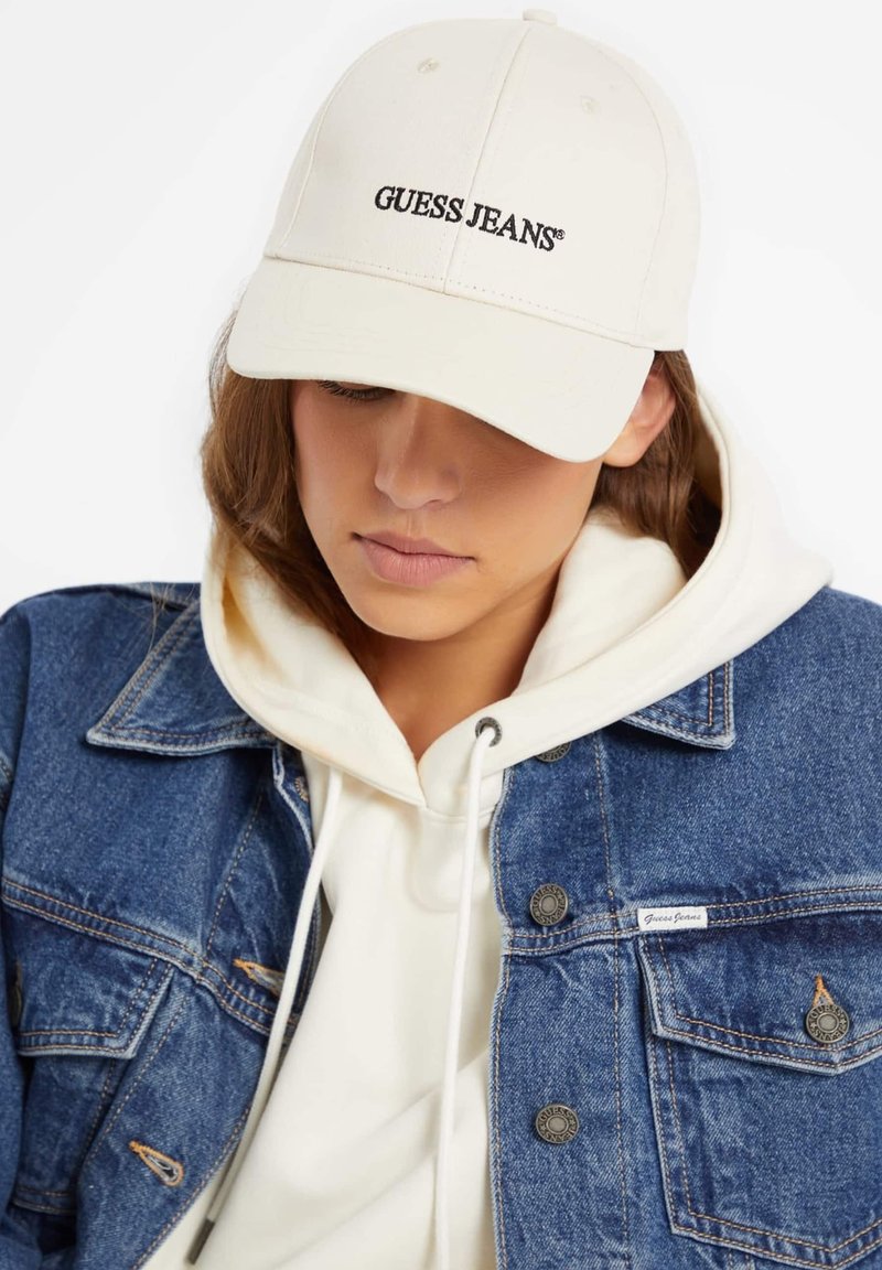 Cappello da baseball crema con logo "GUESS JEANS", abbinato a un hoodie bianco e a una giacca di jeans blu con dettagli di bottoni e tasche.