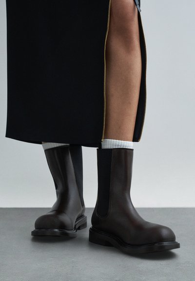 Persoană purtând ghete scurte din piele maro închis cu șosete albe și o fustă neagră cu o fermoar auriu, cu o deschizătură care lasă să se vadă piciorul.