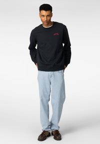 Marine bomullssweatshirt med rød "HEAVY CHOICE" brodering, kombinert med lyseblå denimjeans og brune sko. Modell står med hendene i lommene.