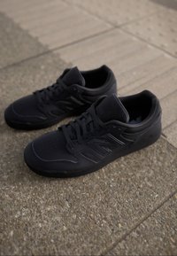 Pár černých tenisek New Balance s tkaničkami, umístěný na texturovaném betoně a dlažbě venku.