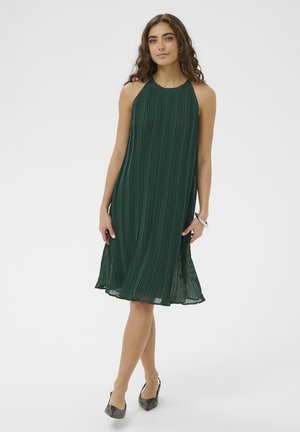 Kobieta o długich, kręconych włosach, ubrana w ciemnozieloną, plisowaną, bezrękawnikową sukienkę midi i ciemne szpilki w szpic, stojąca ze skrzyżowanymi nogami.