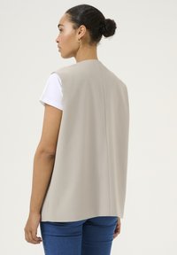 Sleeveless beige garment with a structured design, made of smooth fabric. Features a back seam and worn over a white shirt.