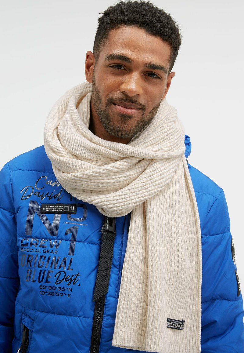 Blue puffer jacket with black branding and zippers, layered with a cream ribbed scarf; the scarf is wrapped around the neck.