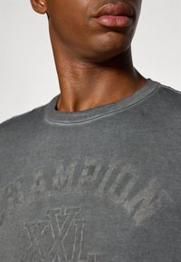 Camiseta de algodón gris con cuello redondo, que presenta un logo "CHAMPION" en tono sobre tono y el detalle "XXL", destacando una textura suave.