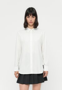 Camisa branca de manga longa com colarinho abotoado, textura suave, corte reto e bainha com fenda lateral, acompanhada por uma saia pregueada preta.