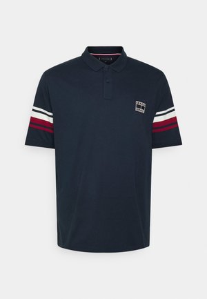 Polo bleu marine à manches courtes avec col, patte à deux boutons, bandes blanches et rouges sur les manches, et petit logo carré sur le côté gauche de la poitrine.