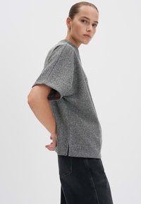 Junge Frau mit slicked-back Haar, die ein lockeres graues, strukturiertes kurzärmliges Oberteil und einen schwarzen Jeansrock trägt, seitlich vor einem weißen Hintergrund posiert.