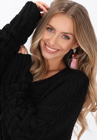 Černý pletený svetr s texturovaným designem, širokými rukávy a hlubokým výstřihem. Doplněno o náušnici s růžovým třásní a vlnitými blond vlasy.