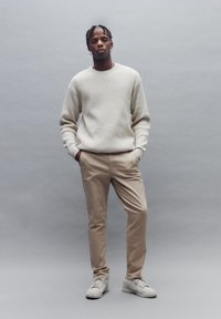 Light beige knitted sweater and beige pants worn with white sneakers, standing against a gray background, hands in pockets.