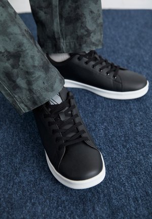 Person trägt schwarze flache Sneakers mit weißen Sohlen und schwarzen Schnürsenkeln und steht auf einem blau carpeted Boden.