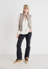 Gilet imbottito beige sopra un maglione bianco con bottoni di perla, jeans neri con fessure laterali e sneakers beige con bordo in pelliccia.