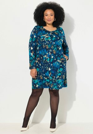Ein blaues und türkisfarbenes gemustertes Kleid mit langen Ärmeln, knielangem Saum und rundem Ausschnitt, kombiniert mit schwarzen Strumpfhosen und beigefarbenen Absätzen.