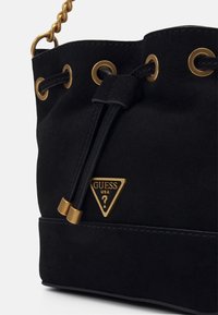 Bolso tipo cubo de ante negro con ojales en tono dorado, cierre de cordón y placa de logo triangular. Superficie texturizada con correa de cadena.